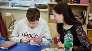 Puisis tumšiem matiem, pelēkā džemperī ar uzrakstu "Liepāja" sēž pie galda un griež rozā papīra lapu. Viņa darbošanos vēros jauna sieviete tumšiem gariem matiem un melnā blūzē.