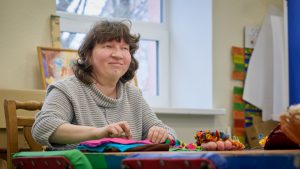 Sieviete tumšiem cirtainiem matiem pelēkā džemperī, sēž pie galda un nodarbojas ar rokdarbiem, izmantojot krāsainus auduma un dzijas materiālus.