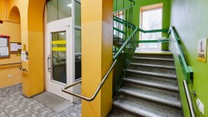 Telpas interjers. Kāpņu telpa ar koridori. Sienas oranžā un zaļā krāsā. Kreisajā pusē kāpmes ar margām, labajā lifts,.