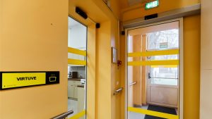 Koridoris oranžā krāsā ar divām stiklotām durvīm. Kreisajā pusē plāksnīte ar uzrakstuj "Virtuve"