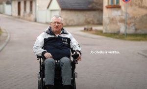 Kungs īsiem sirmiem matiem, tumšā jakā pārvietojas pa klusu, bruģētu vecpilsētas ielu elektriskajā ratiņkrēslā